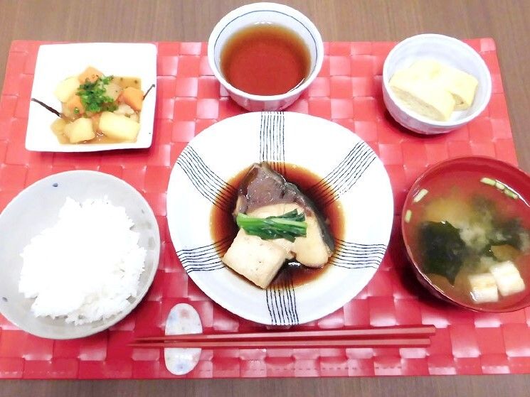 魚の煮つけに白いご飯、みそ汁、野菜の煮物などが並んだ赤いランチョンマット。