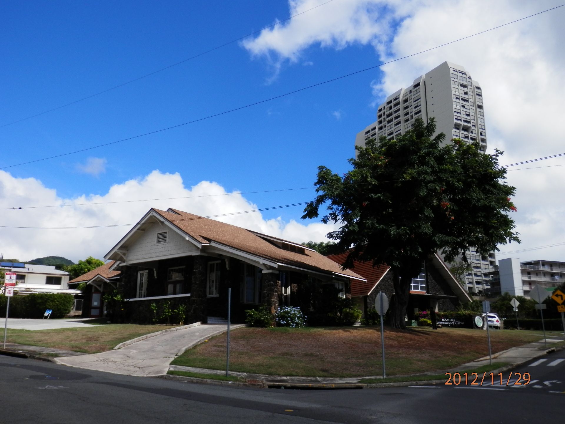 青い空の下に建っているブラウンの屋根の平屋建ての建物。周りには緑が多くあり、奥には白いビルがある。