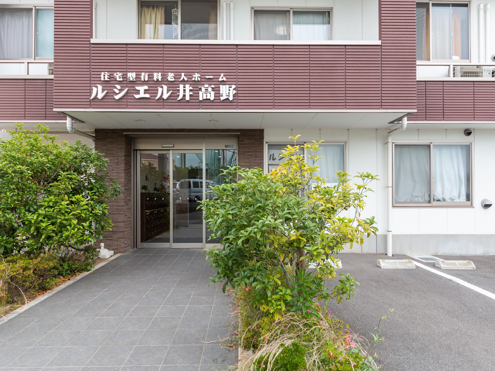 日当たりのいい室内で笑顔で過ごしている車いすの高齢女性とピンクのエプロンを付けた若い女性スタッフ。