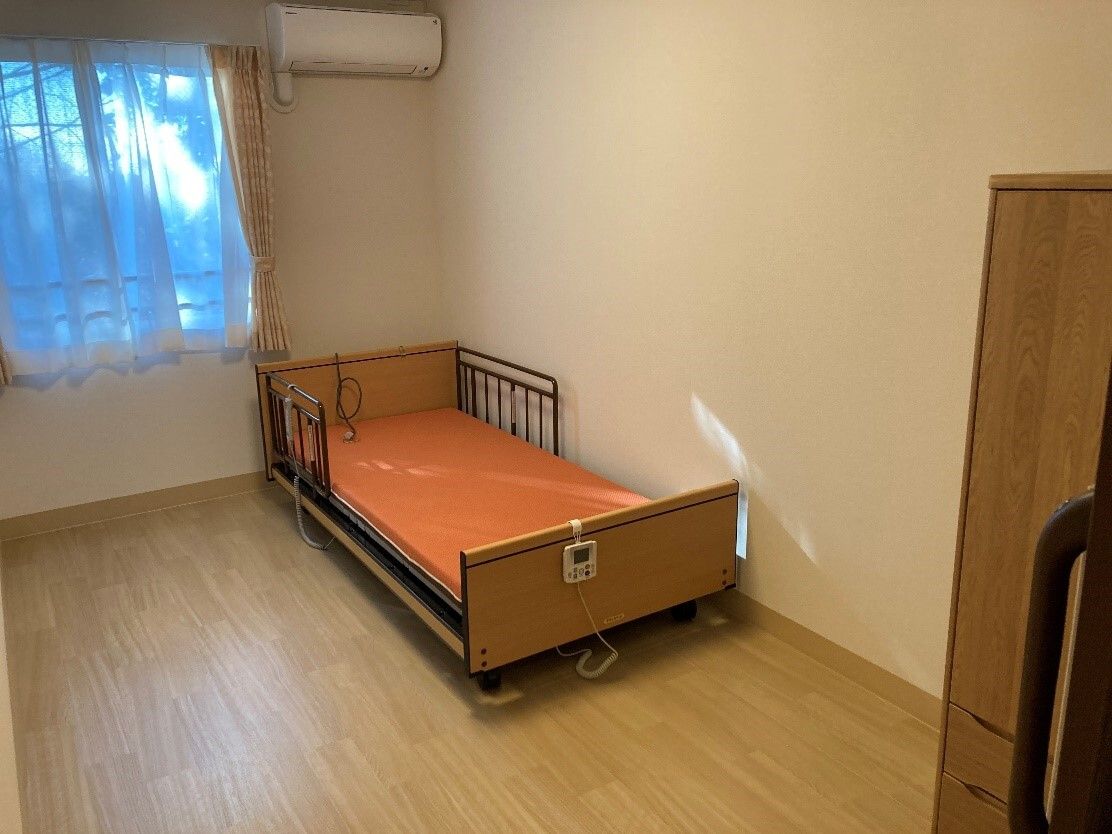 レースカーテンとカーテンが付いた窓があるフローリングの部屋にはエアコンや介護ベッドがあり、ドアはスライド式。