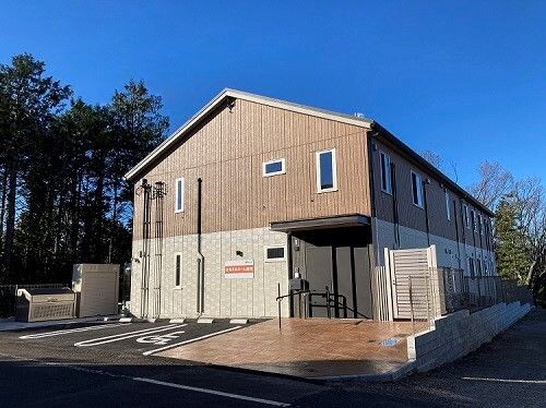 ２階建てのブラウン系の建物の前には駐車場があり、玄関周りには手すりがある。周りには自然も多く残っている。