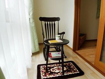 レースカーテンとカーテンが付いた広いサンルームが付いた部屋。サンルームにはロッキングチェアが置かれている。