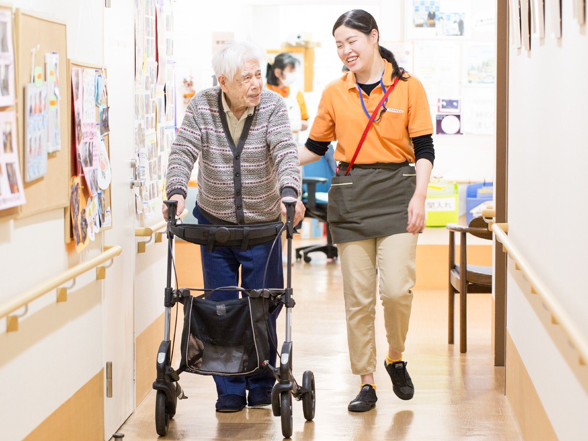 歩行器を利用しながらまっすぐな廊下を歩いている高齢男性に笑顔で話かけている女性スタッフ。