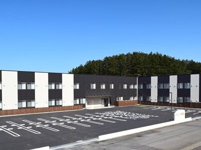 大きな山が後ろにあり、手前には広い駐車場が付いたエル字型の2階建ての建物があり、モノトーンの外壁。