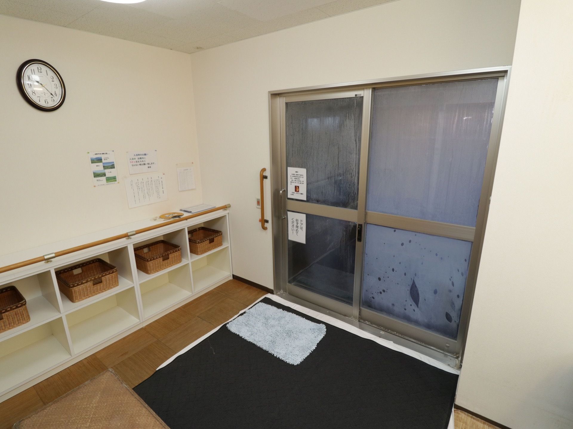 白を基調とした衛生的な脱衣所である。棚は２段で低く使いやすく、手すりが完備されている。滑らないようにマットが何枚も敷かれている。