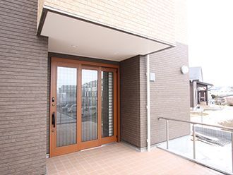 屋根が付いた玄関にはスライド式のドアが設置されていて、手すりもある。周辺にも建物がある。