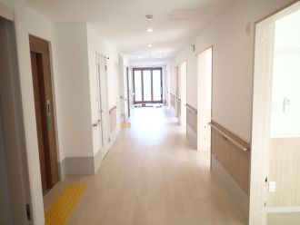 手すりが付いたまっすぐな廊下の突き当りには玄関のドアがあり、左側のエレベーターの前には点字ブロックがある。