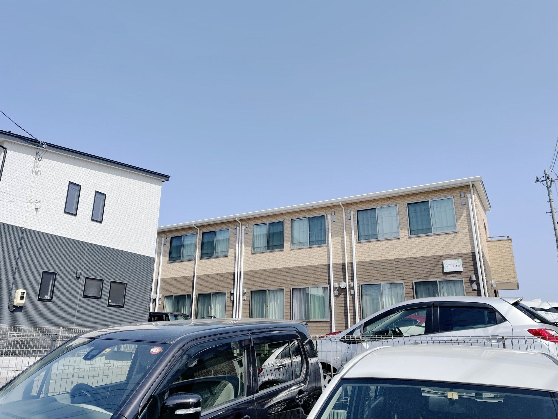 ベージュとブラウンの2階建ての建物には屋根が付き、玄関には手すりがあり、広い駐車場もある。