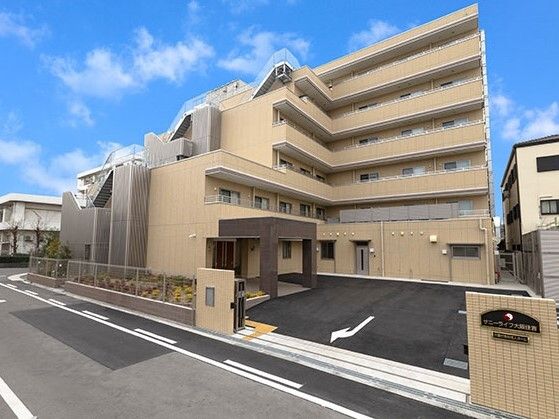 階段のようなデザインの建物はベージュ系の外壁で敷地内には駐車場があり、赤や黄色の花が植えられた花壇もある。