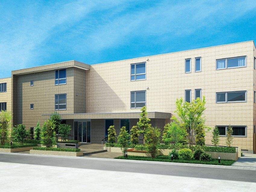 横に長い3階建ての建物はベージュ系の外壁で、玄関には屋根があり、手すり付きの階段とスロープが付いている。