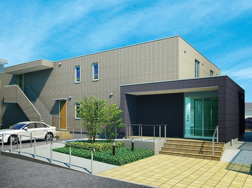グレー系の２階建ての建物の前には駐車場があり、玄関には大きな屋根が付き、階段とスロープがある。