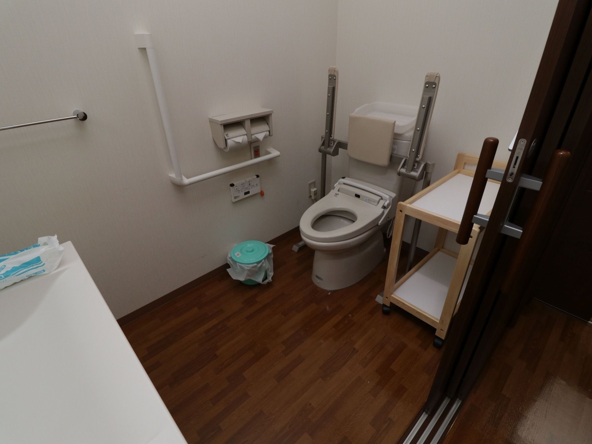 スペースの広いトイレである。Ｌ型手すりや背面付きアームレストが完備されている。汚物入れやワゴンが置かれている。
