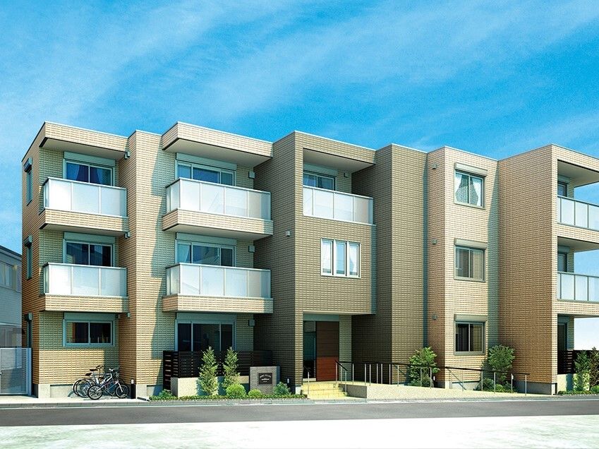 ブラウン系の建物は3階建てで玄関には手すり付きの階段とスロープがあり、建物の前には駐輪場がある。