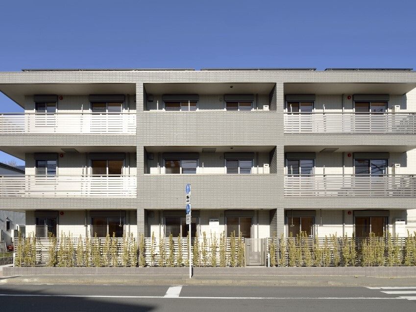 グレーの外観の3階建ての建物の周りには草木が植えられ、横に長いデザイン。各フロアにベランダがある。