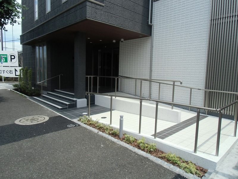 ベージュとダークグレーの4階建ての建物は草木に囲まれ、玄関には手すり付きのスロープと階段が付いている。
