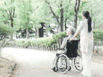 自然豊かな場所を車いすで散歩している高齢男性とそれを補助しながら話している女性スタッフ