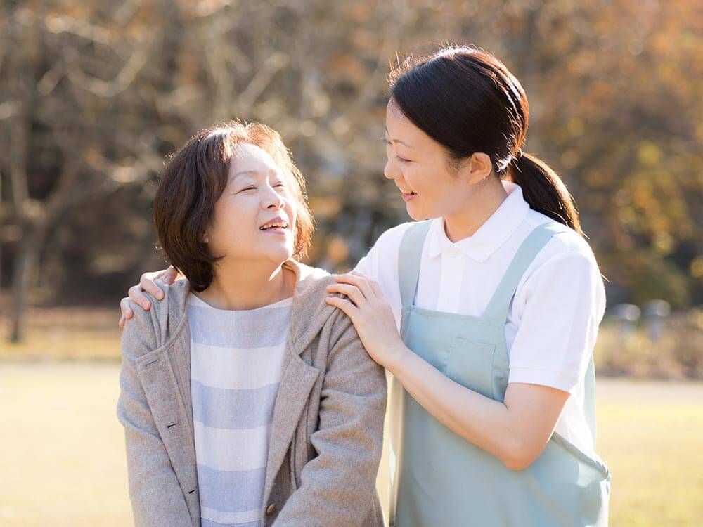 原っぱのような場所を散歩している高齢女性のそばにブルーのエプロンを付けた若い女性スタッフが付き添っている。