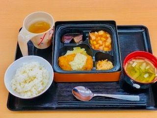 四角い弁当風の入れ物におかずが盛られ、みそ汁とお茶、ご飯が添えられている。トレーにはスプーンもある。