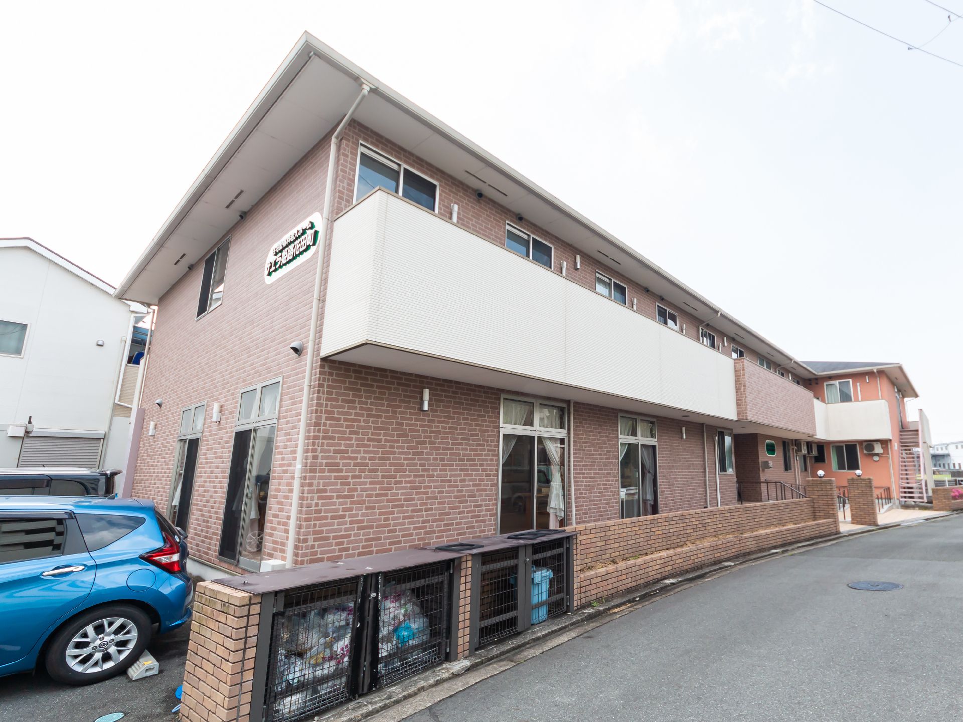 2階建ての建物はブラウン系で２階部分にはベランダが付き、玄関には手すり付きの階段とスロープもある。