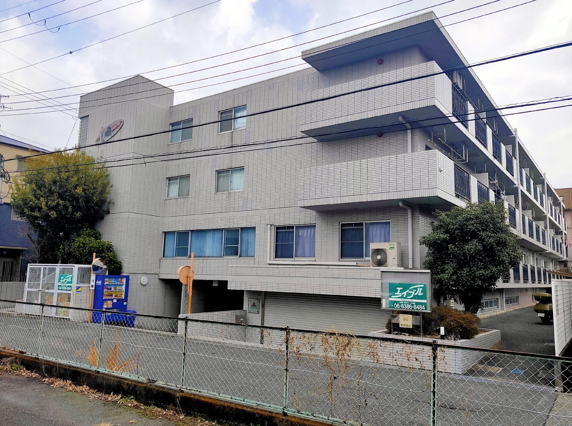 各フロアにベランダがあり、重厚感のある造りの３階建ての建物。敷地内にはブルーの自動販売機も置かれている。