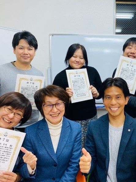 眼鏡をかけた講師ような女性を中心に症状を持った男女が4名、持っていない男性が一人笑顔で写真撮影に応じている。