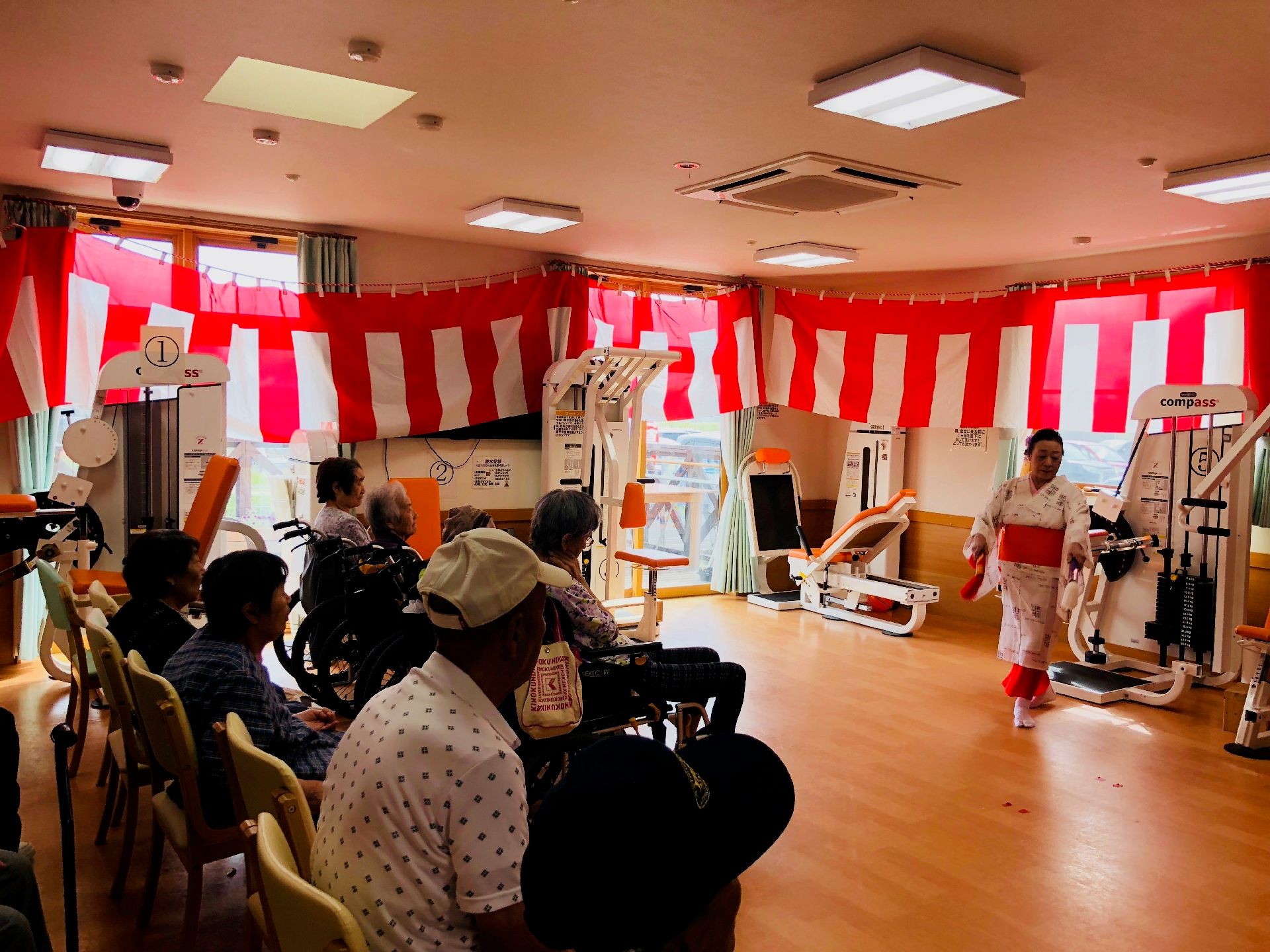 着物を着た女性の踊りを見ている高齢者たち。部屋の壁には紅白の垂れ幕が飾られている。