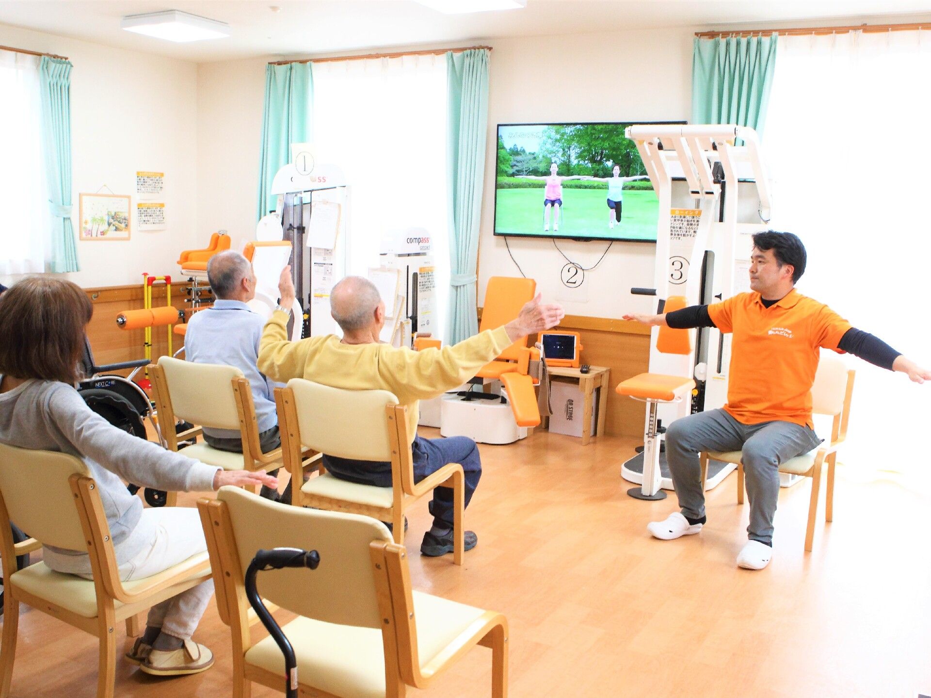 オレンジのポロシャツを着た男性スタッフが前に座り、テレビと一緒に高齢者たちの体操の指導をしている。