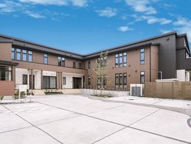 エル字型の建物はダークブラウンの外壁で敷地内の駐車場には車が止まっている。周りには草木が植えられている。