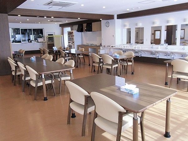 厨房や自動販売機、手洗い場が見えて、ベージュの椅子と茶色いテーブルが並んでいる食堂