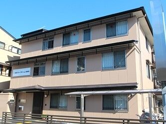 3階建てのベージュ系の建物の各フロアに屋根が付いていて、玄関には大き目の屋根が付いている。