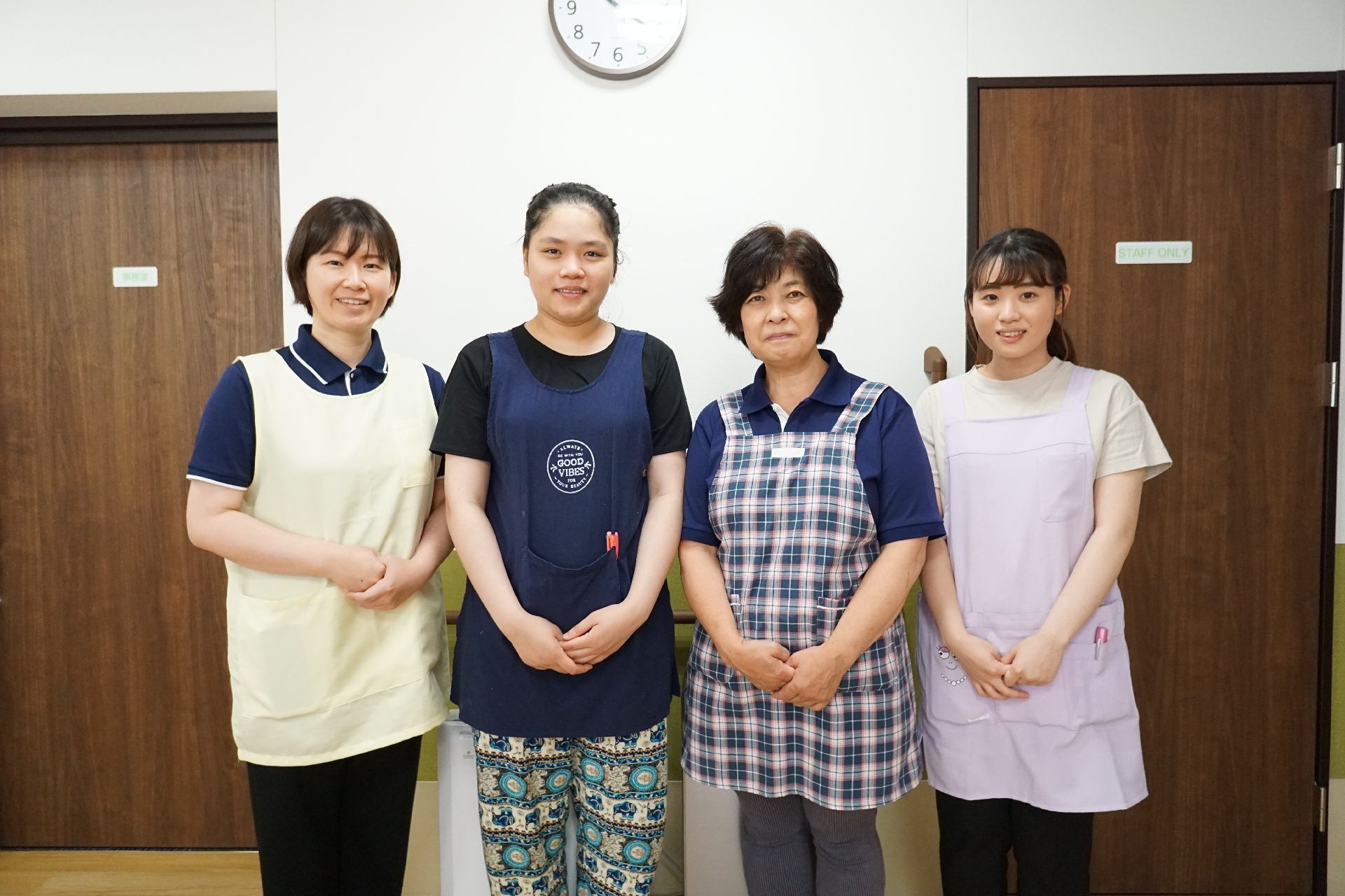 緑の多い屋外で笑顔で過ごしている車いすの高齢女性に寄り添っているエプロンを付けた若い女性スタッフ。