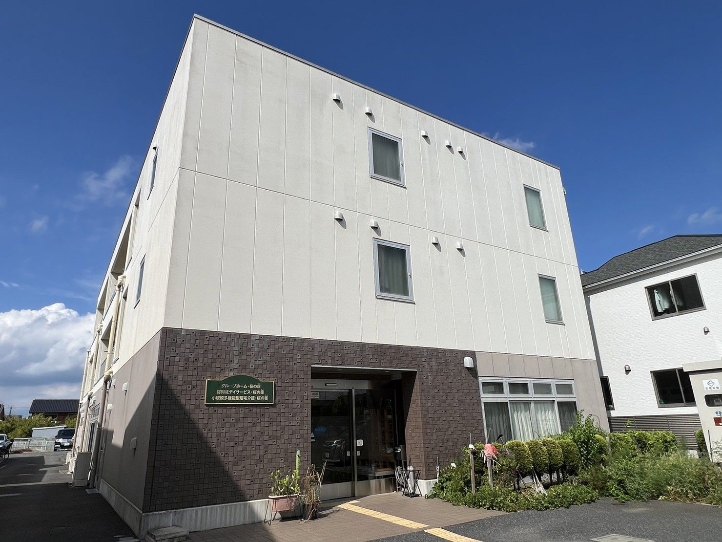 3階建ての建物の1階部分はブラウン、2階と3階は白で、敷地内駐車場には点字ブロックも敷かれている。