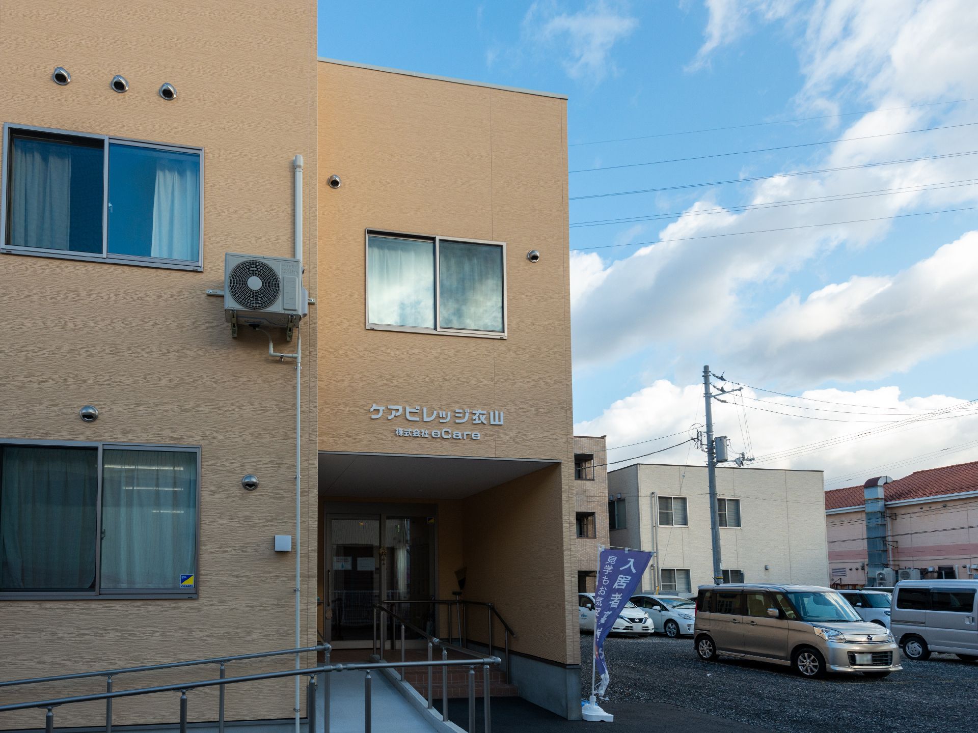 車いすの高齢男性のそばにはゆったりとした服を着た高齢女性が建っていて、男性の肩に手を置き笑いかけている。