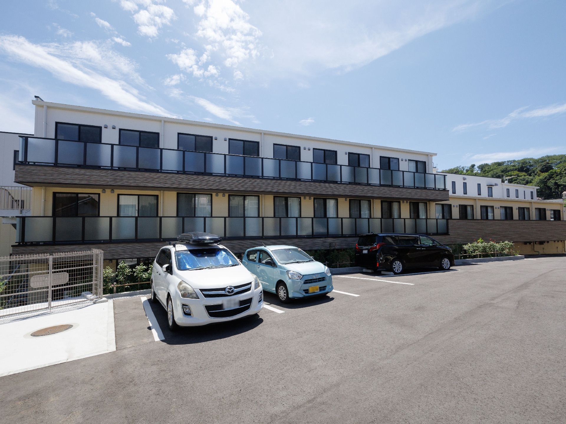 バルコニーが付いた白と黄色の外観の建物の側面に設けられた駐車スペースに止まる２台の車