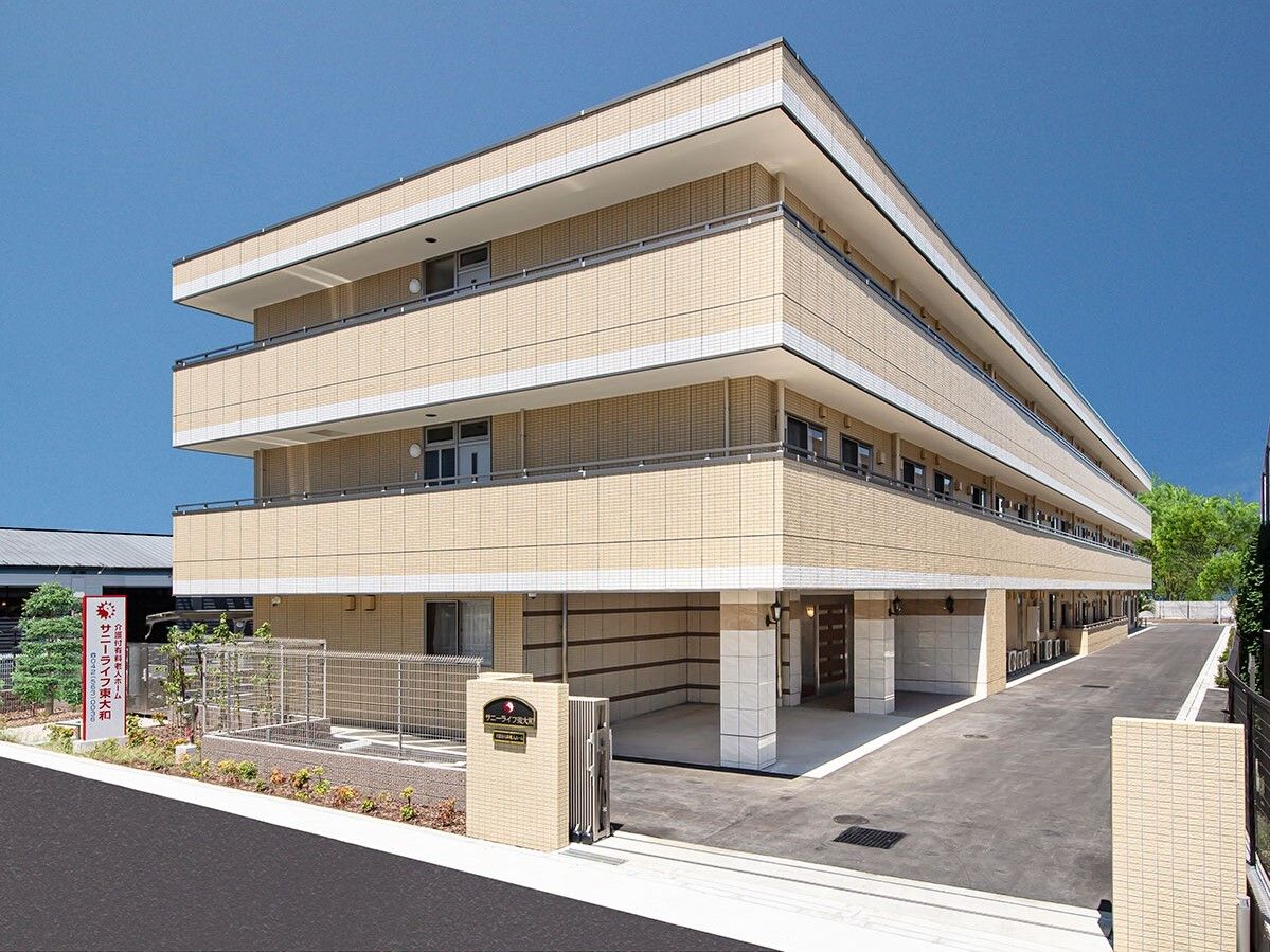 各フロアにベランダがあるベージュ系の３階建ての建物は横に長く、周りには緑も多い。