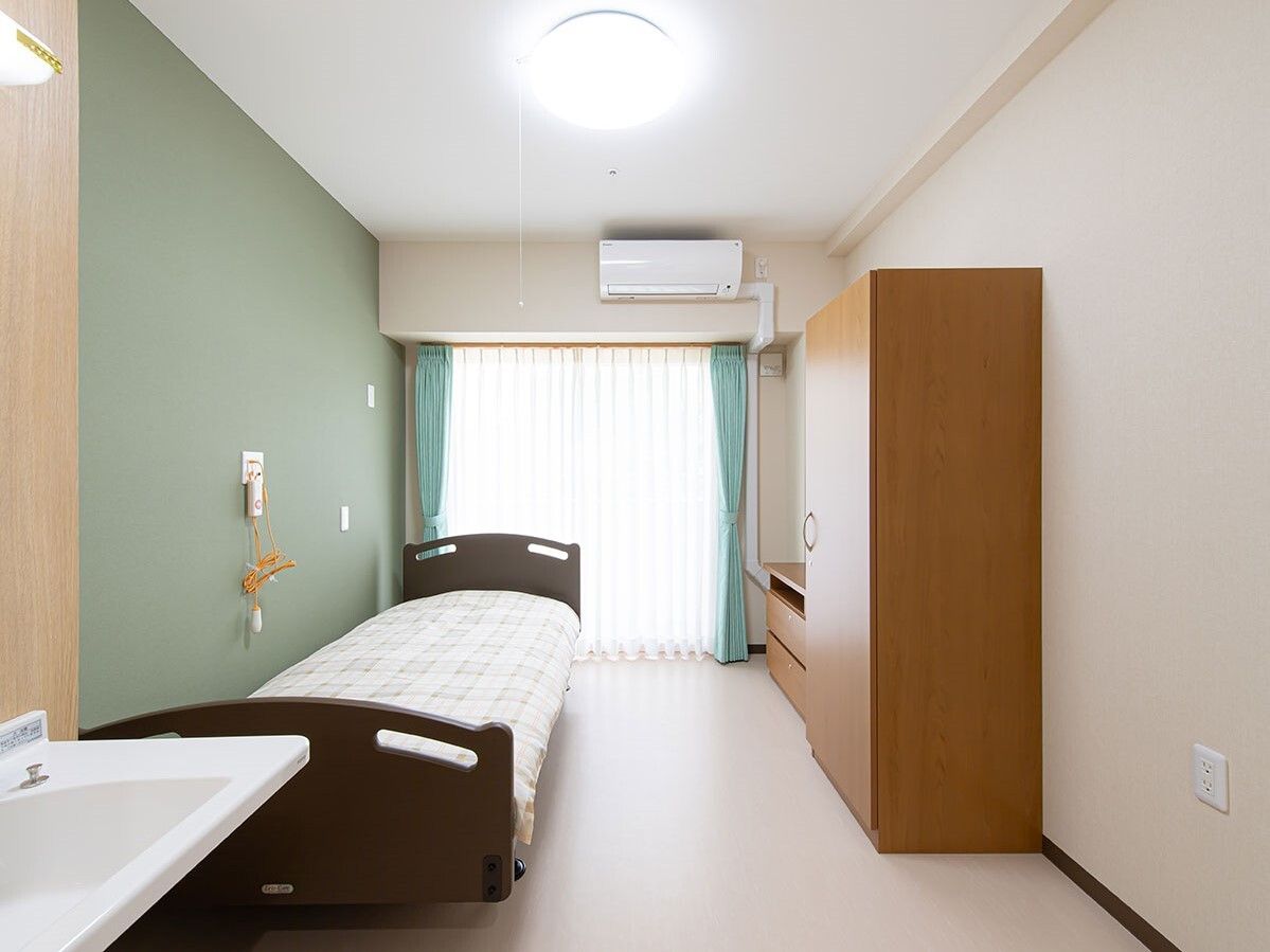 ベッドサイドには緊急通報装置が付いた個室の部屋には寝具付きのベッドが置かれ、収納家具もある。