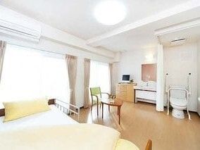 カーテンで開け閉めするタイプのトイレや車いす対応の洗面所、介護ベッド、エアコン、テーブルセットなどがある部屋。