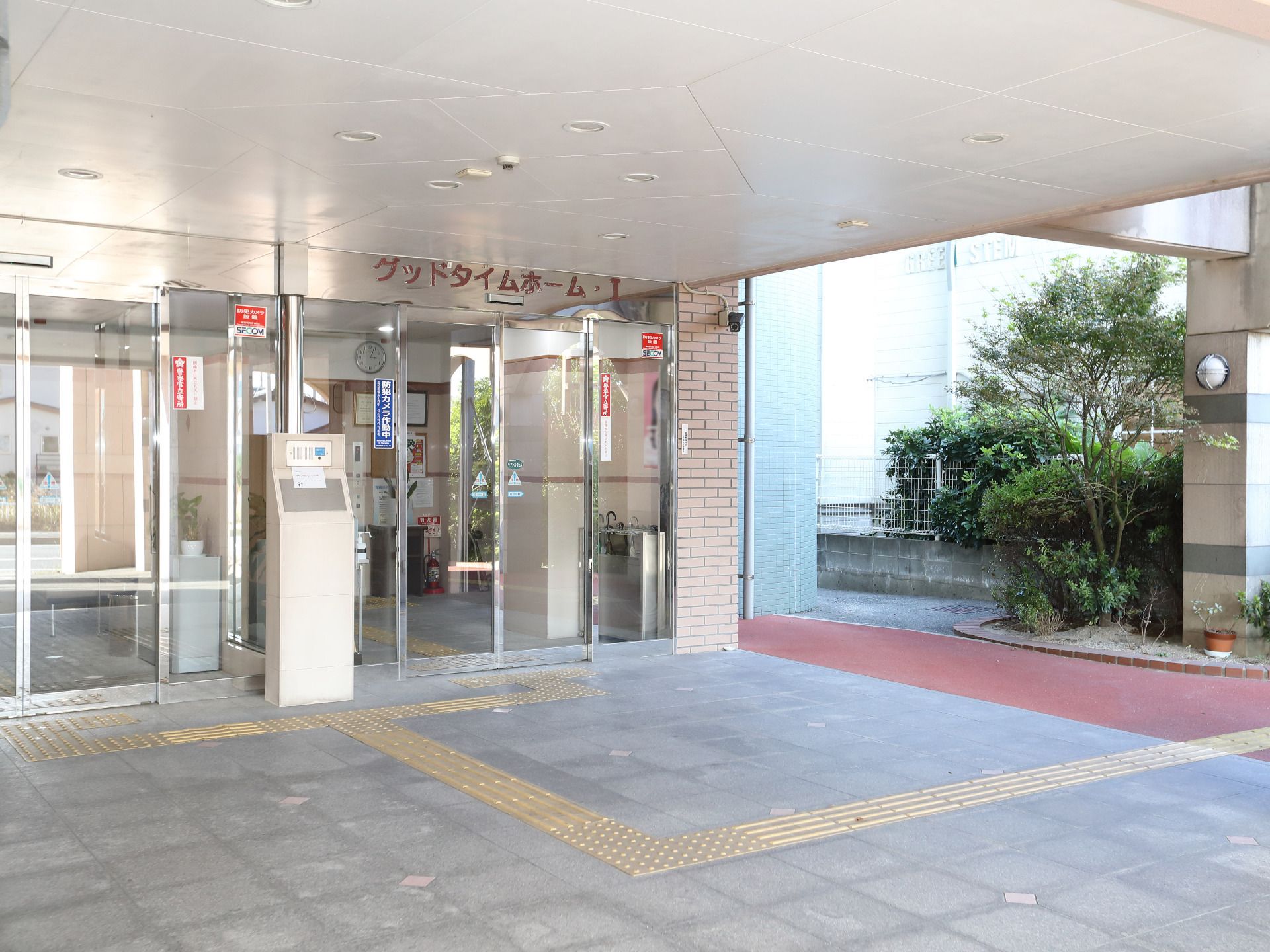 カーディガンを着た高齢女性がピンクのエプロンを付けた女性スタッフと並んで寄り添い、笑顔で過ごしている。