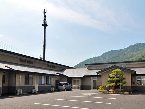 後方に山があり、手前には広い駐車場があり、平屋建ての建物には三角屋根が付いている。