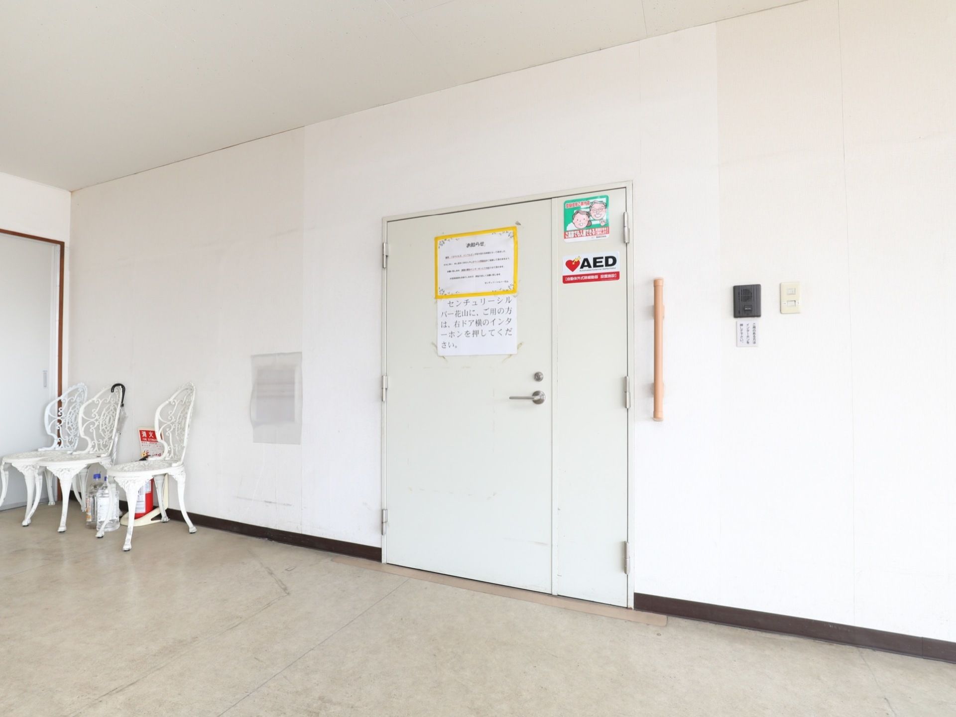 天井や壁が白色で統一されている部屋で、正面に鉄製の扉がある。壁にインターフォンが付いている。扉の左には、椅子が3脚ある。