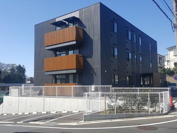 フェンスに囲まれた3階建ての建物はダークグレーの外壁で日当たりのいい場所に建っている。駐車場もある。