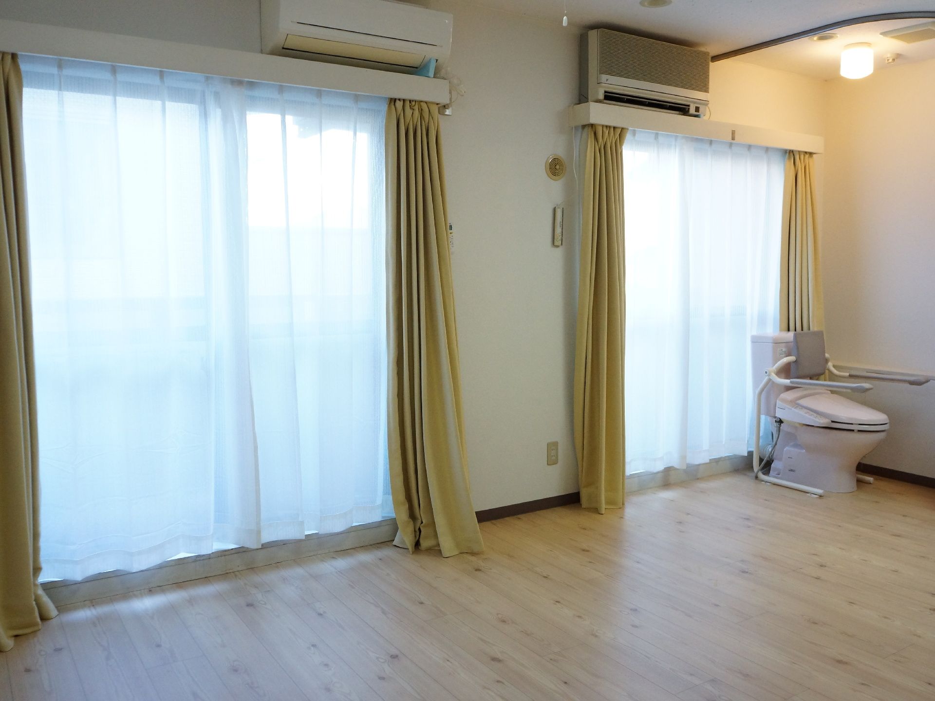 木目調の床となっている個室は窓が2つ写っており、奥の方にはトイレが設置されている。トイレの上では照明が点いている。