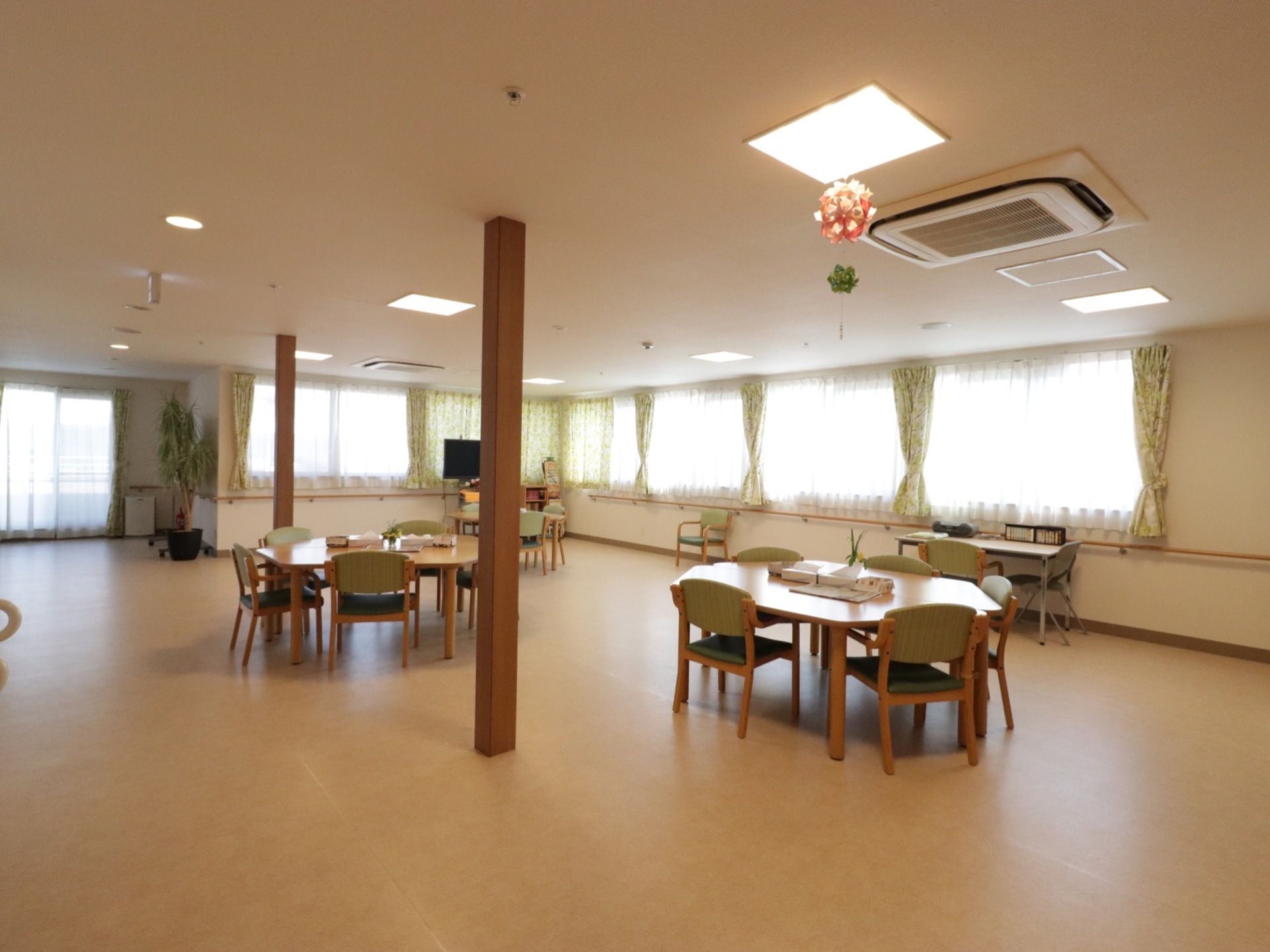 外に面した壁に、腰高窓が連なっている。窓際にテレビがある。中央付近に柱がある。テーブルセットが3セット程度ある。