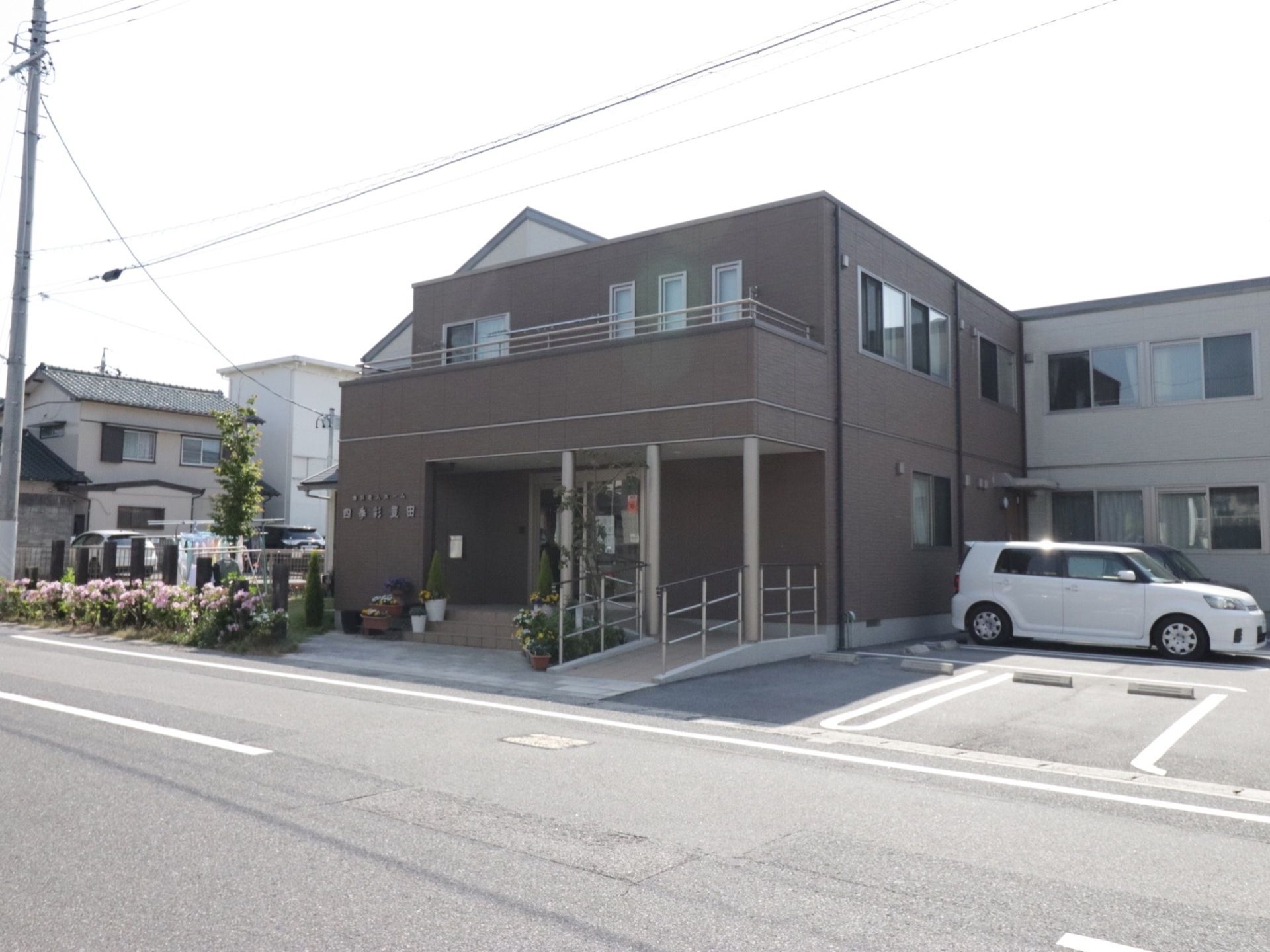 建物は2階建てで住宅街にあり道路に面している。隣に数台停められる駐車場がある。駐車場から玄関へは歩道に出てから向かう。