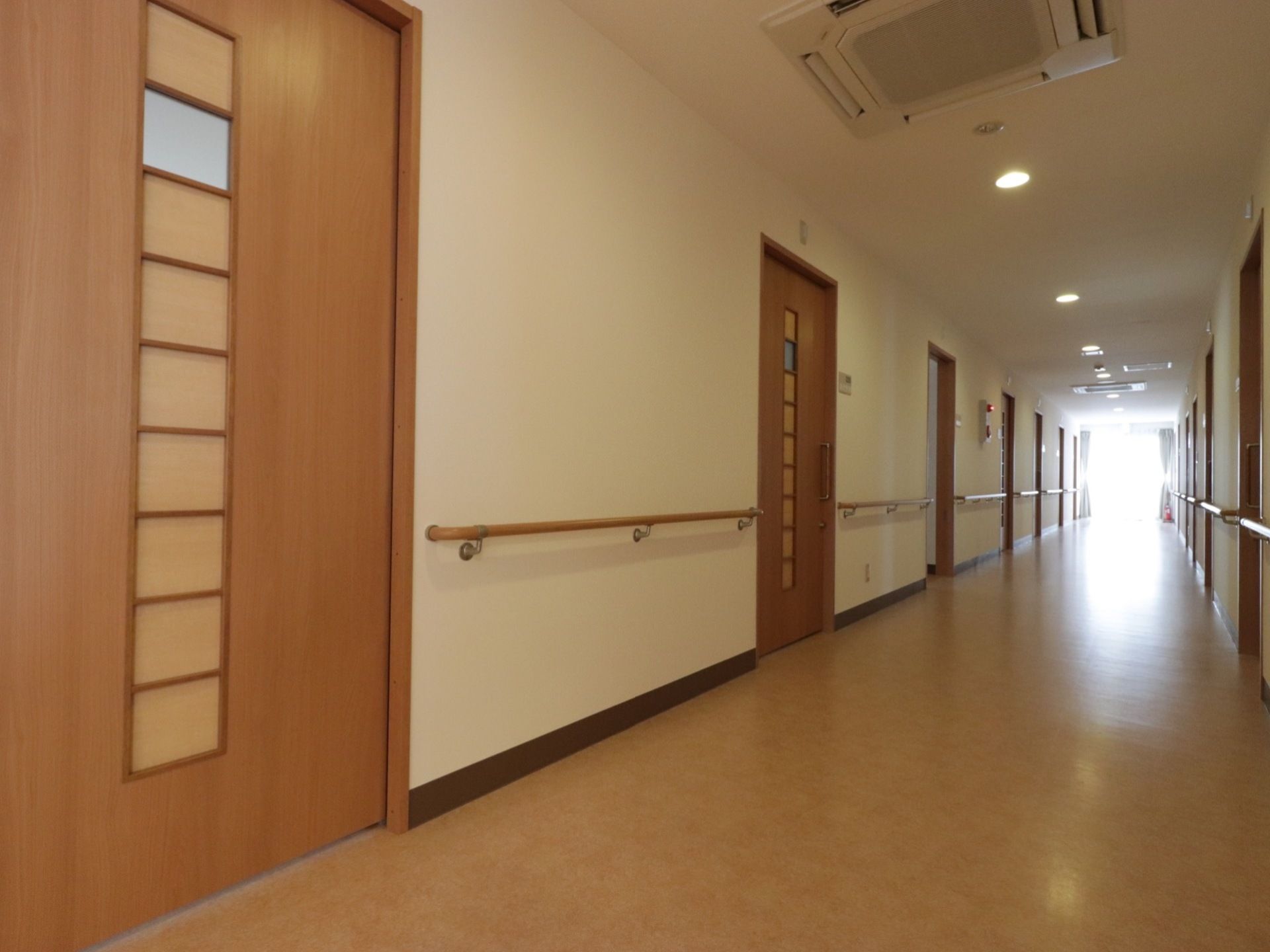廊下の両側に居室のドアが続いている。また壁には手すりが設置してある。木製のドアは中央にデザインを施している。