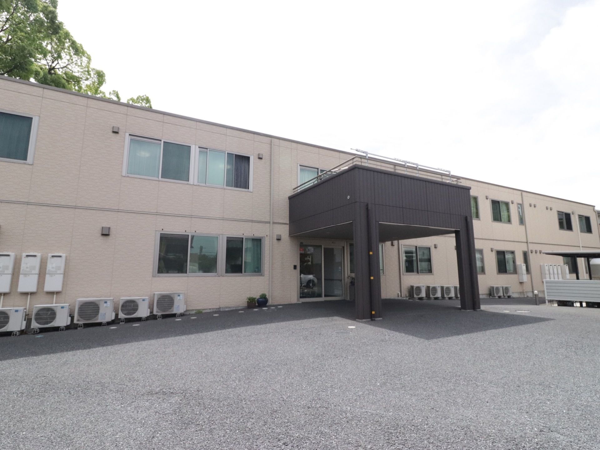 ２階建てのベージュの建物である。建物の前面には、室外機が並んでいる。玄関前には屋根があり、車は屋根の下に入れる。