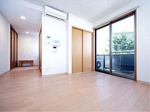 白い壁にフローリングの室内。ベランダに繋がる窓からは木の緑が見える。壁にはクーラーが設置されている。