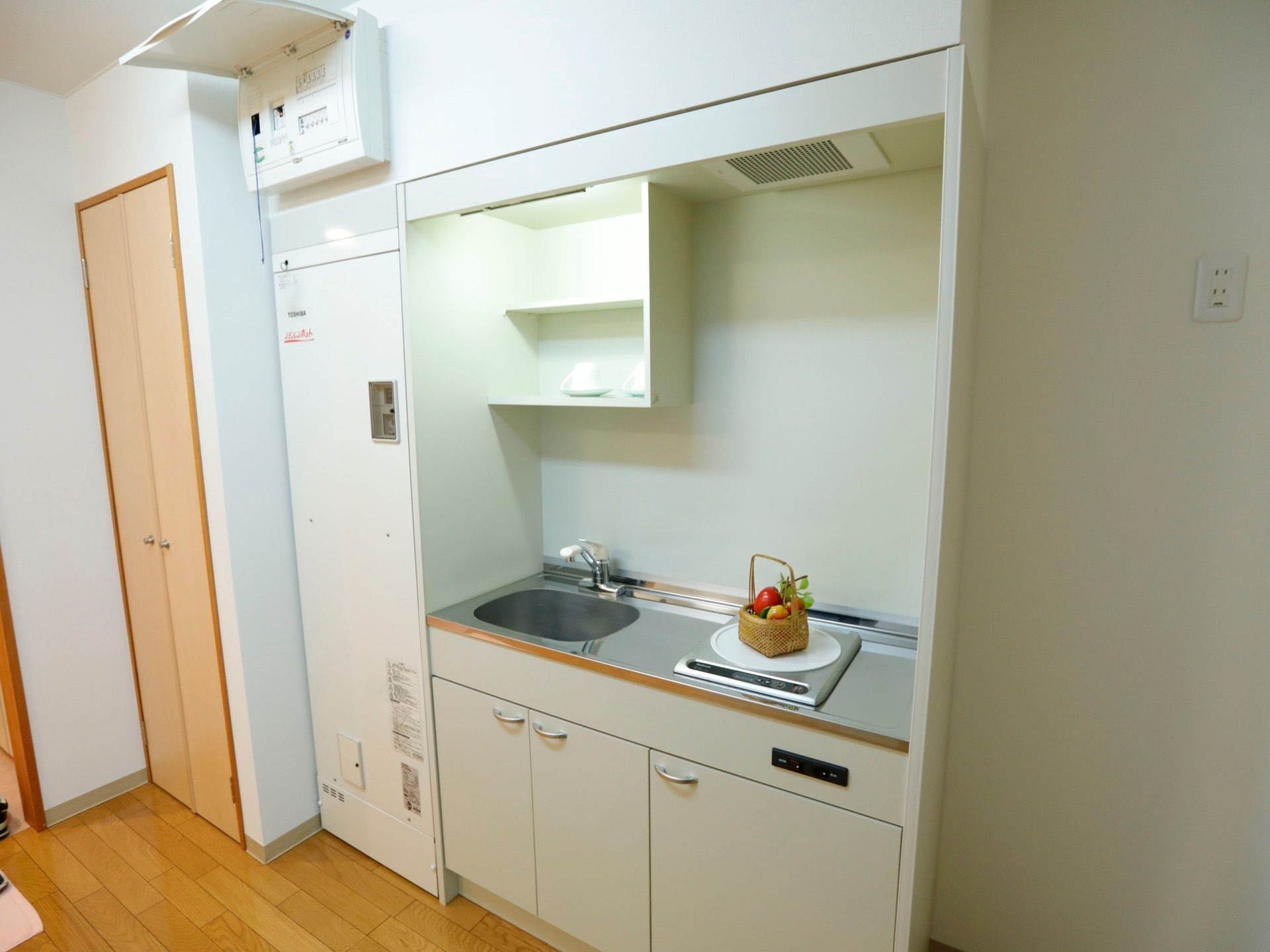右側に電磁調理器、左側に流し台、その上に収納スペースという造りのミニキッチンである。小さいカゴの中に、赤い果物が入っている。