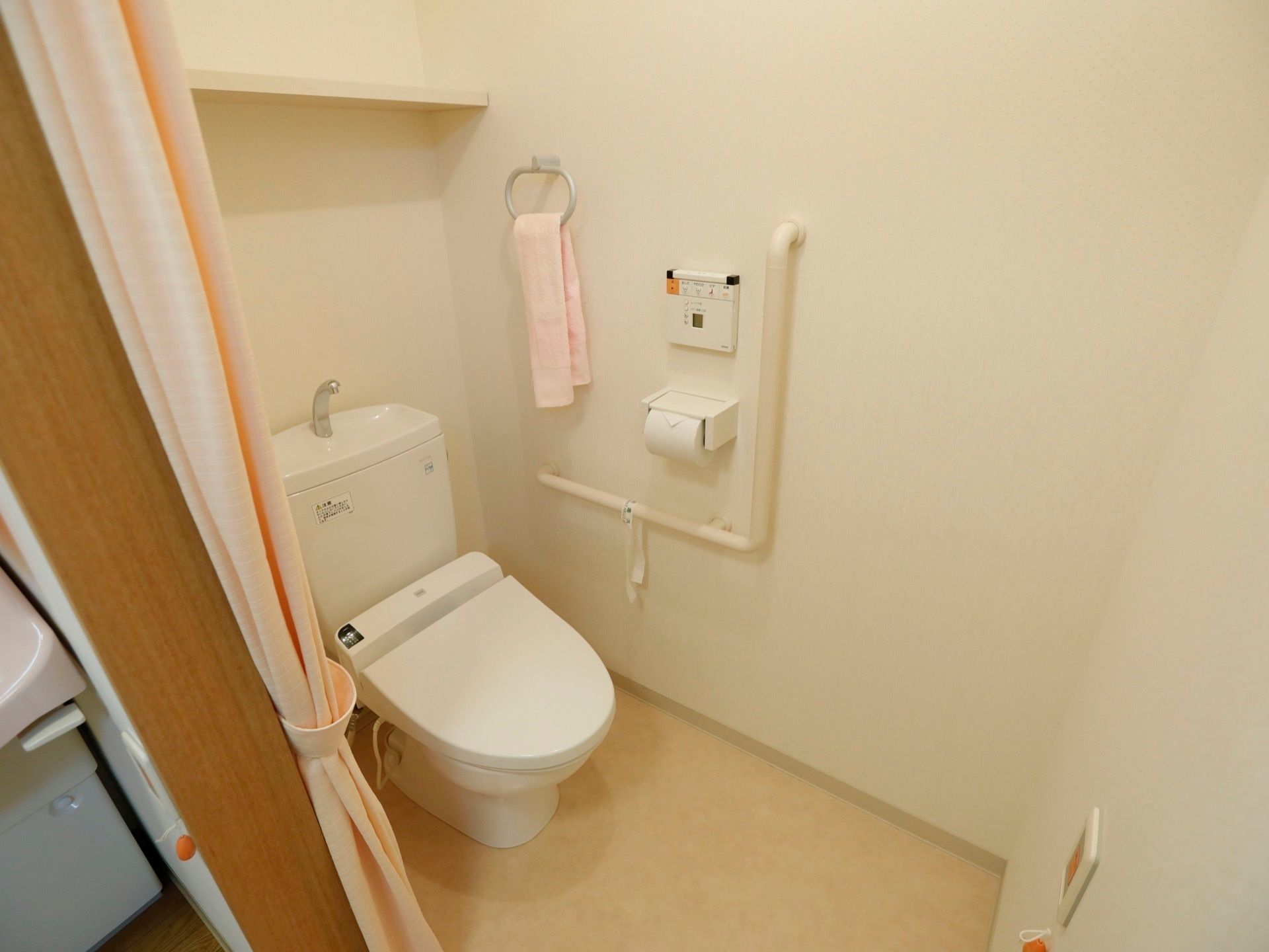 トイレの右側の壁には、L字型の手すりと、ペーパーホルダーが取り付けられている。その横には、淡いピンク色のタオルが掛けてある。