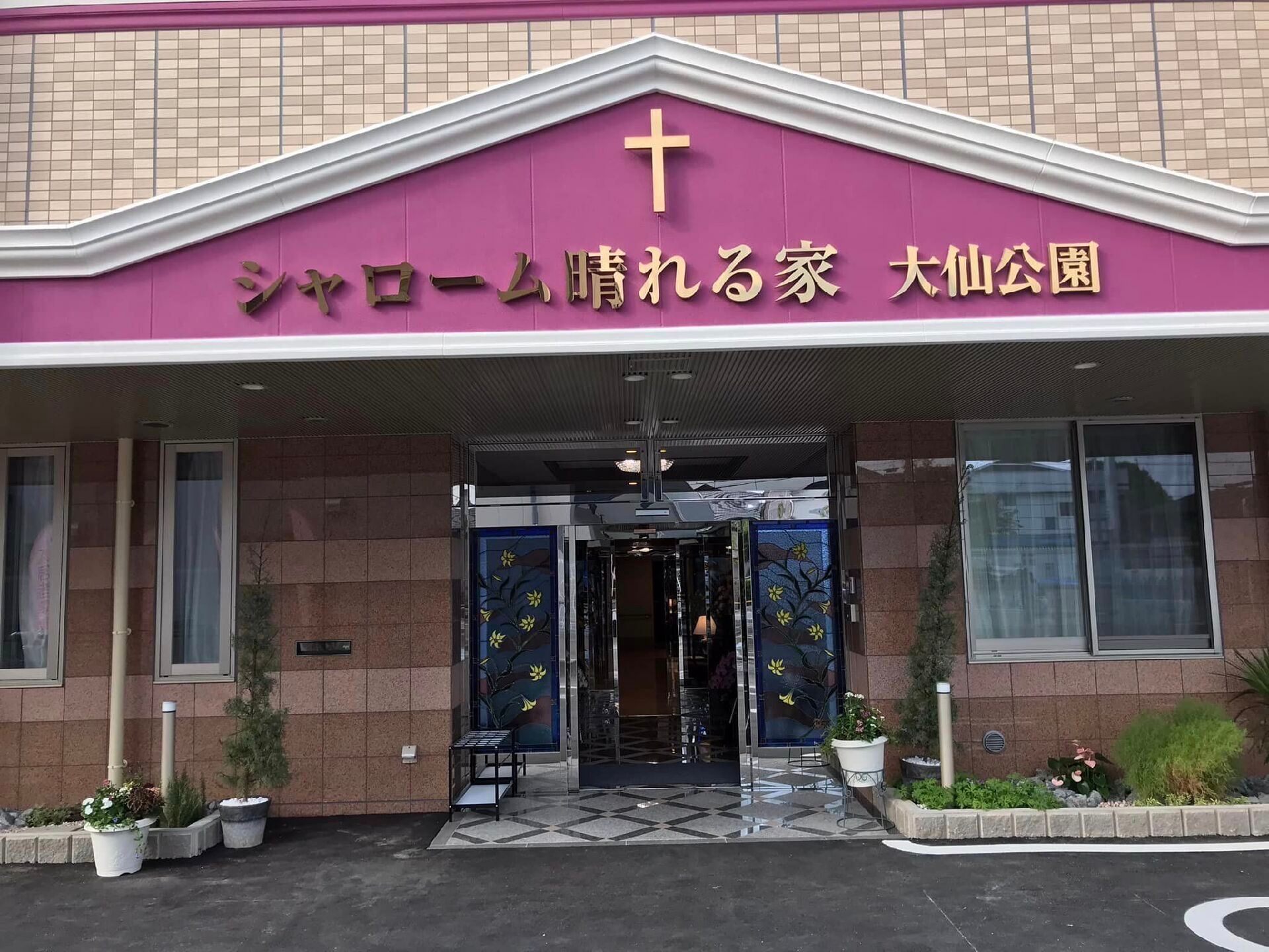 窓がたくさんある４階建ての建物の周りには草木が植えられていてブラウン系でまとめられている。